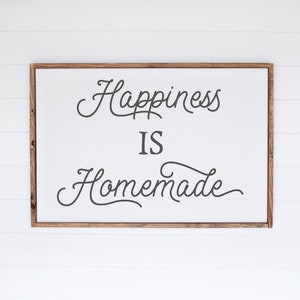May include: A framed sign with the text "Happiness is Homemade" in a cursive font. The sign is on a white wood background.
