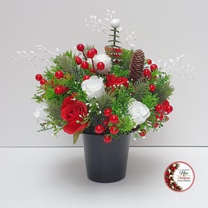 May include: A floral arrangement in a black pot featuring red and white roses, green pine needles, red berries, a brown pinecone, and white decorative branches. A circular logo in the bottom right corner reads All About Nice Christmas Grace Flowers.