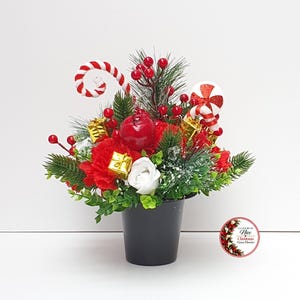 May include: A festive holiday arrangement in a black pot featuring red and white flowers, green pine branches, red berries, miniature gold gift boxes, and candy cane accents. A small circular logo with the text Nice Christmas Green Flowers is visible.