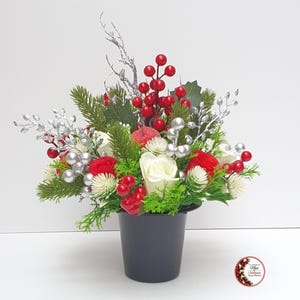 May include: A floral arrangement in a dark grey pot featuring red and white roses, green pine needles, bright red berries, silver-coloured decorative branches, and small white thistle-like flowers. A small circular logo is visible in the bottom right.
