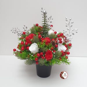 May include: A floral arrangement in a black pot featuring red and white roses, green pine needles, bright red berries, a small central pine tree, and silver decorative branches. A circular logo with the text Nee Christmas Green Flowers is visible.