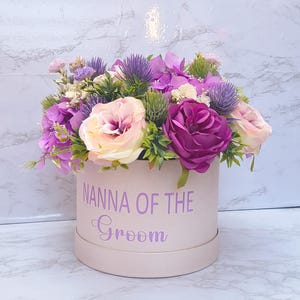 May include: A round, light pink gift box featuring the purple text "NANNA OF THE Groom" on the front, filled with an arrangement of artificial flowers including purple roses, pale pink roses, purple thistles, and green foliage.