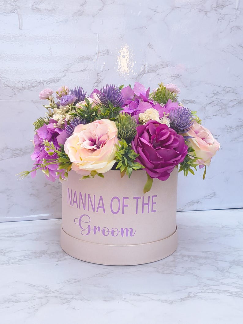 May include: A round, light pink gift box featuring the purple text "NANNA OF THE Groom" on the front, filled with an arrangement of artificial flowers including purple roses, pale pink roses, purple thistles, and green foliage.