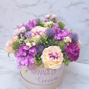 May include: A round beige gift box containing an arrangement of artificial flowers in shades of purple, lavender, and cream. The box features the purple text NANNA OF THE Groom printed on the front. The floral display includes roses, hydrangeas, and thistles.