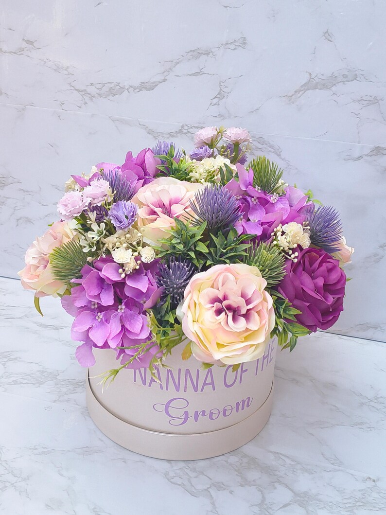May include: A round beige gift box containing an arrangement of artificial flowers in shades of purple, lavender, and cream. The box features the purple text NANNA OF THE Groom printed on the front. The floral display includes roses, hydrangeas, and thistles.