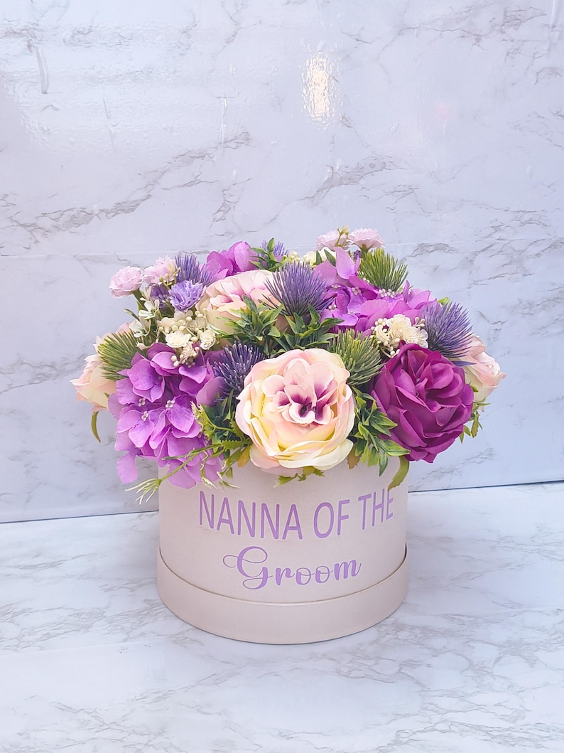 May include: A round, light pink gift box featuring the purple text "NANNA OF THE GROOM" in a mix of block and script fonts. The box is filled with an arrangement of artificial flowers, including purple hydrangeas, light pink roses, and thistle.