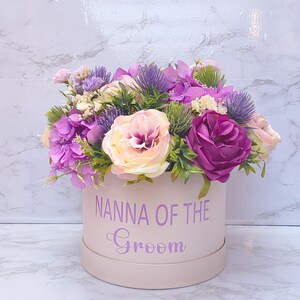 May include: A round, light pink gift box featuring the purple text "NANNA OF THE Groom" on the front. The box is filled with an arrangement of artificial flowers, including purple hydrangeas, deep magenta roses, pale pink roses, and spiky thistle.