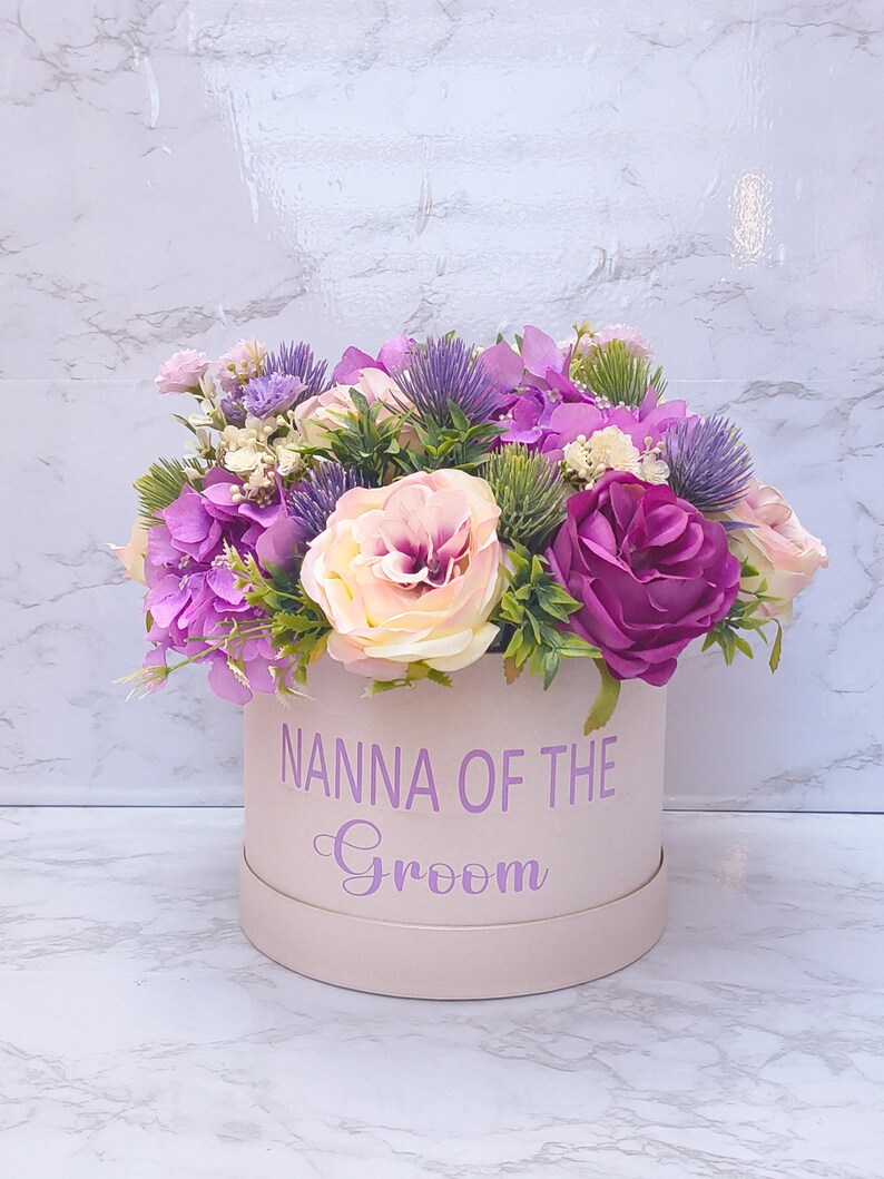 May include: A round, light pink gift box featuring the purple text "NANNA OF THE Groom" on the front. The box is filled with an arrangement of artificial flowers, including purple hydrangeas, deep magenta roses, pale pink roses, and spiky thistle.
