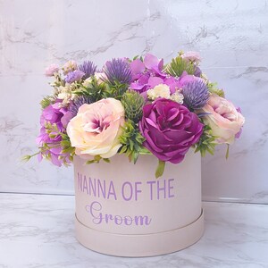 May include: A round, light pink gift box featuring the purple text NANNA OF THE Groom, filled with an arrangement of artificial flowers including purple roses, light pink roses, purple thistle, and small green foliage accents.