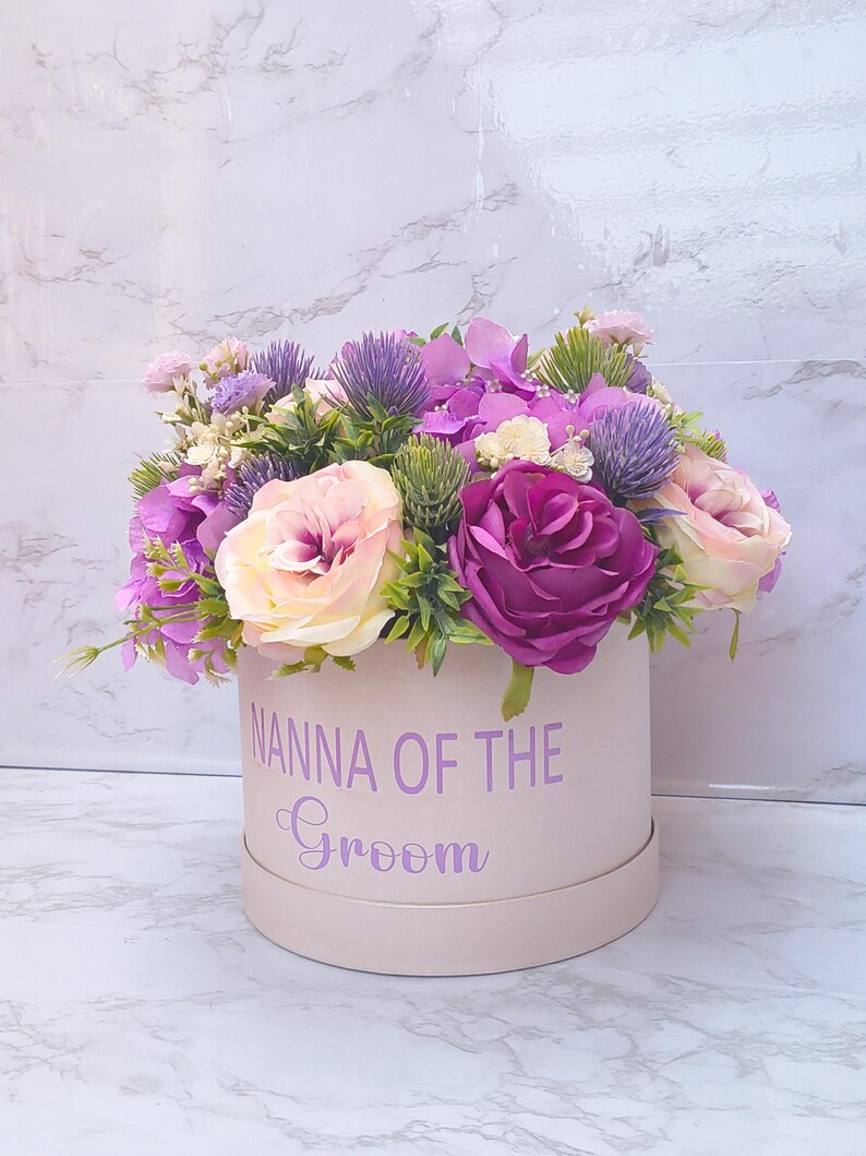 May include: A round, light pink gift box featuring the purple text NANNA OF THE Groom, filled with an arrangement of artificial flowers including purple roses, light pink roses, purple thistle, and small green foliage accents.