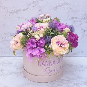 May include: A round, light pink gift box featuring the purple text NANNA OF THE Groom, filled with an arrangement of artificial flowers including light pink roses, vibrant purple hydrangeas, and spiky purple thistle accents.