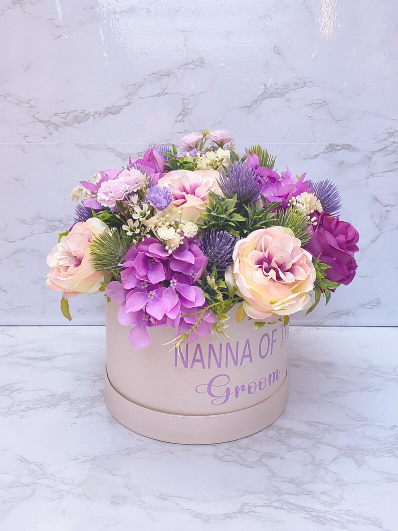 May include: A round, light pink gift box featuring the purple text NANNA OF THE Groom, filled with an arrangement of artificial flowers including light pink roses, vibrant purple hydrangeas, and spiky purple thistle accents.