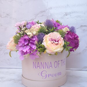 May include: A round, light pink gift box featuring the purple text NANNA OF THE Groom, filled with an arrangement of artificial flowers including purple hydrangeas, light pink roses, purple thistle, and green foliage.