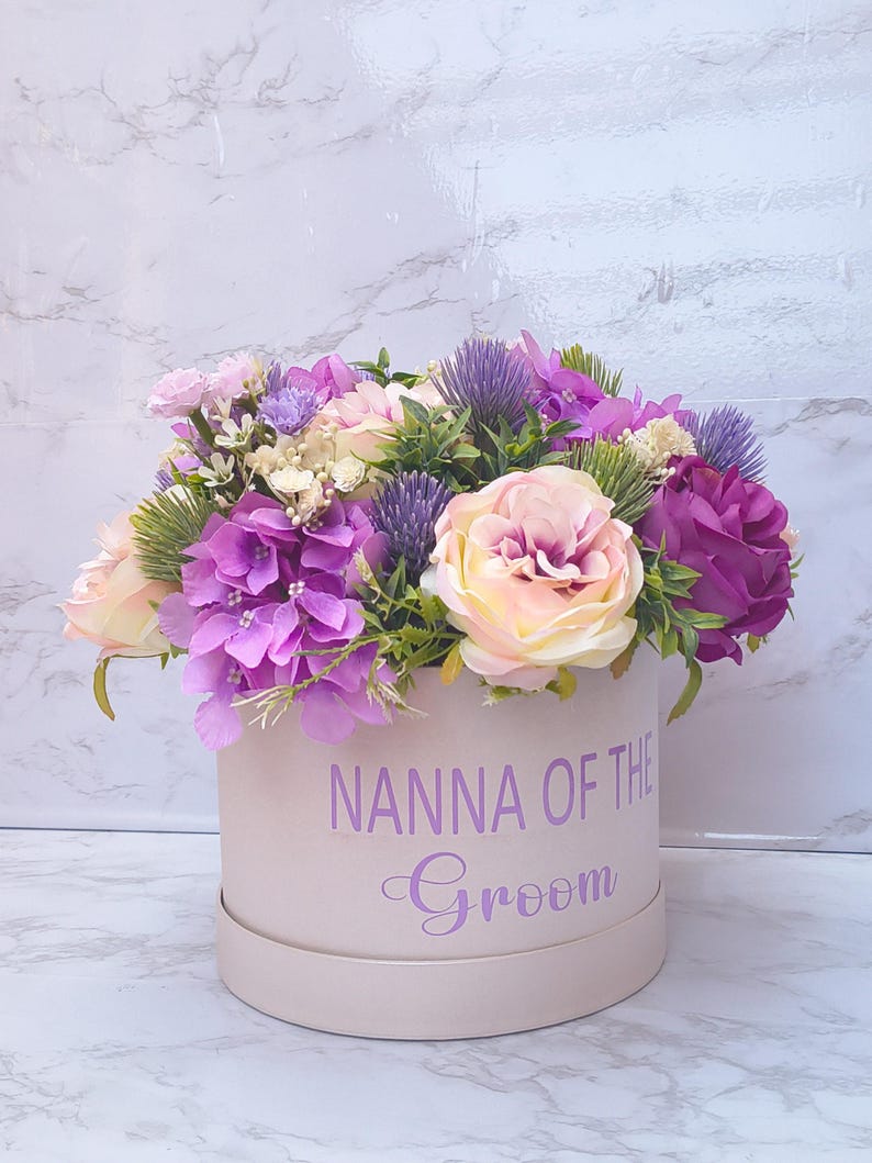May include: A round, light pink gift box featuring the purple text NANNA OF THE Groom, filled with an arrangement of artificial flowers including purple hydrangeas, light pink roses, purple thistle, and green foliage.