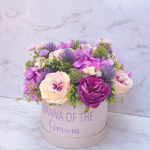 May include: A round light pink gift box featuring the purple text NANNA OF THE GROOM, filled with an arrangement of artificial flowers including purple hydrangeas, deep magenta roses, pale pink roses, and spiky purple thistle accents.