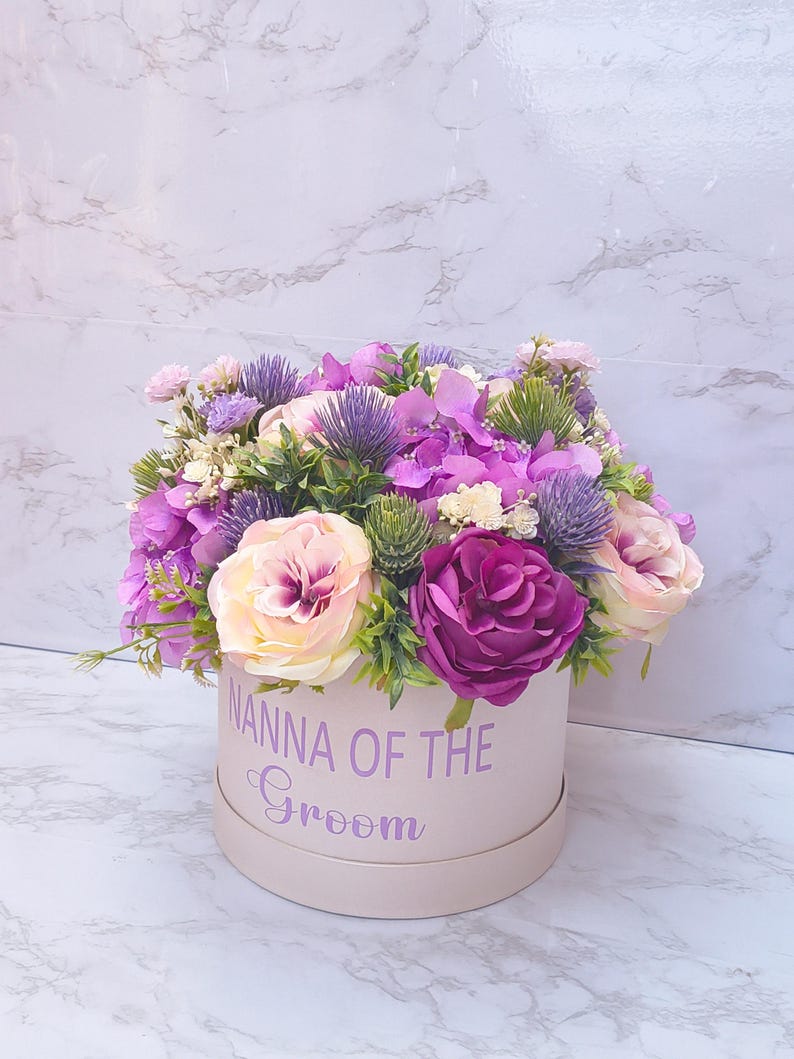 May include: A round light pink gift box featuring the purple text NANNA OF THE GROOM, filled with an arrangement of artificial flowers including purple hydrangeas, deep magenta roses, pale pink roses, and spiky purple thistle accents.