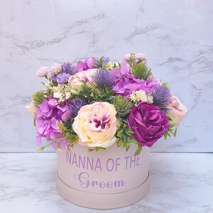 May include: A round light pink gift box featuring the text NANNA OF THE Groom in purple lettering. The box is filled with an arrangement of artificial flowers including purple hydrangeas, cream and dark pink roses, and spiky blue thistle.