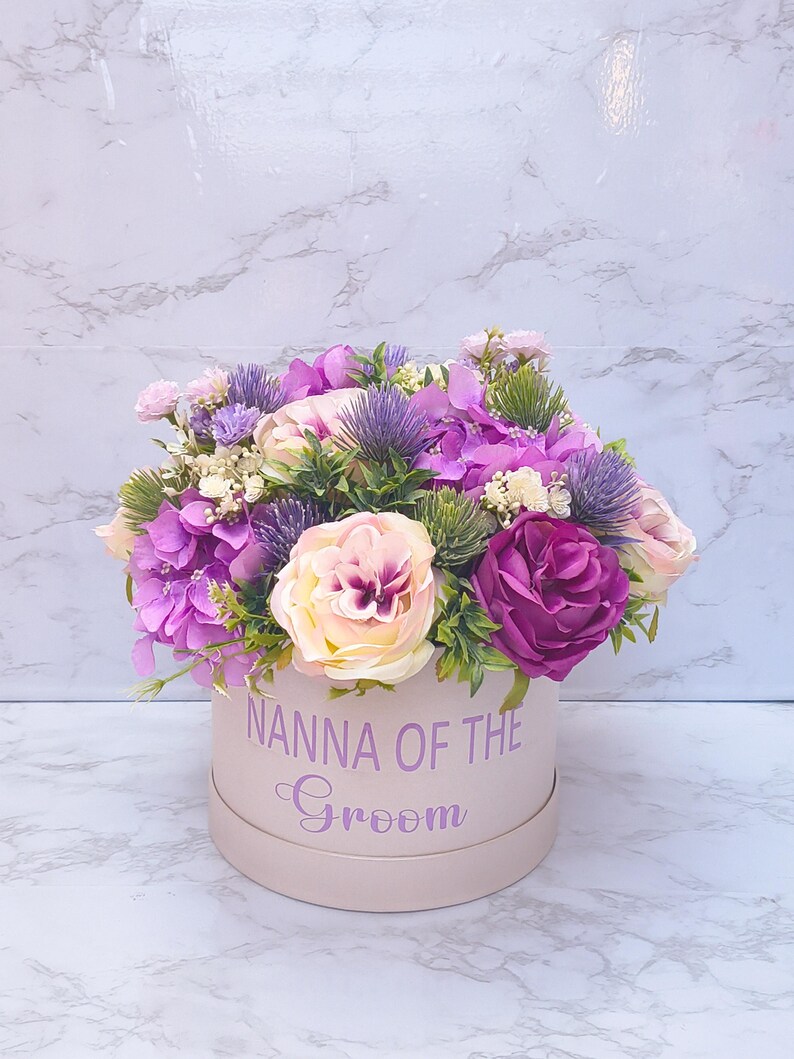 May include: A round light pink gift box featuring the text NANNA OF THE Groom in purple lettering. The box is filled with an arrangement of artificial flowers including purple hydrangeas, cream and dark pink roses, and spiky blue thistle.