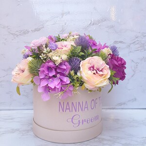 May include: A round, light pink gift box featuring the purple text NANNA OF THE Groom, filled with an arrangement of artificial flowers including light pink roses, vibrant purple hydrangeas, and spiky purple thistle accents.