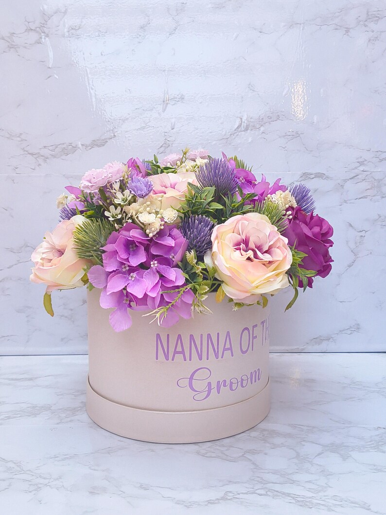 May include: A round, light pink gift box featuring the purple text NANNA OF THE Groom, filled with an arrangement of artificial flowers including light pink roses, vibrant purple hydrangeas, and spiky purple thistle accents.