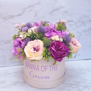 May include: A round light pink gift box with the text NANNA OF THE Groom printed in purple. The box is filled with an arrangement of artificial flowers including purple hydrangeas, cream and deep magenta roses, and spiky purple thistle accents.