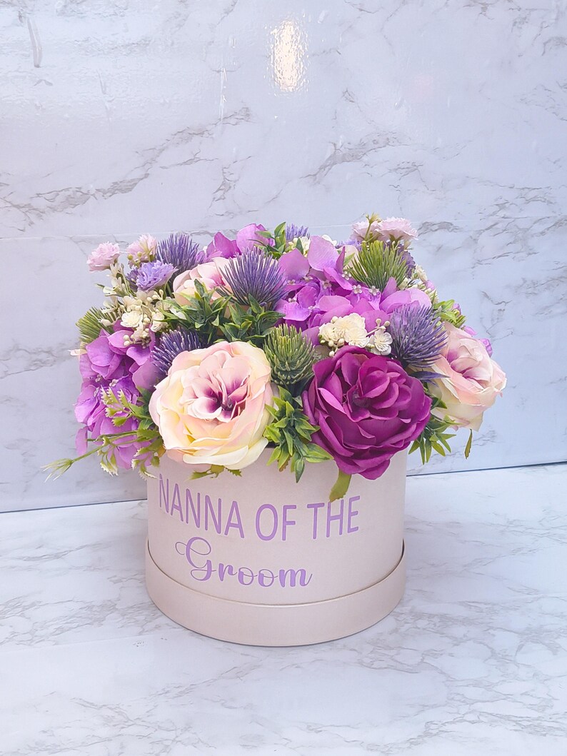 May include: A round light pink gift box with the text NANNA OF THE Groom printed in purple. The box is filled with an arrangement of artificial flowers including purple hydrangeas, cream and deep magenta roses, and spiky purple thistle accents.