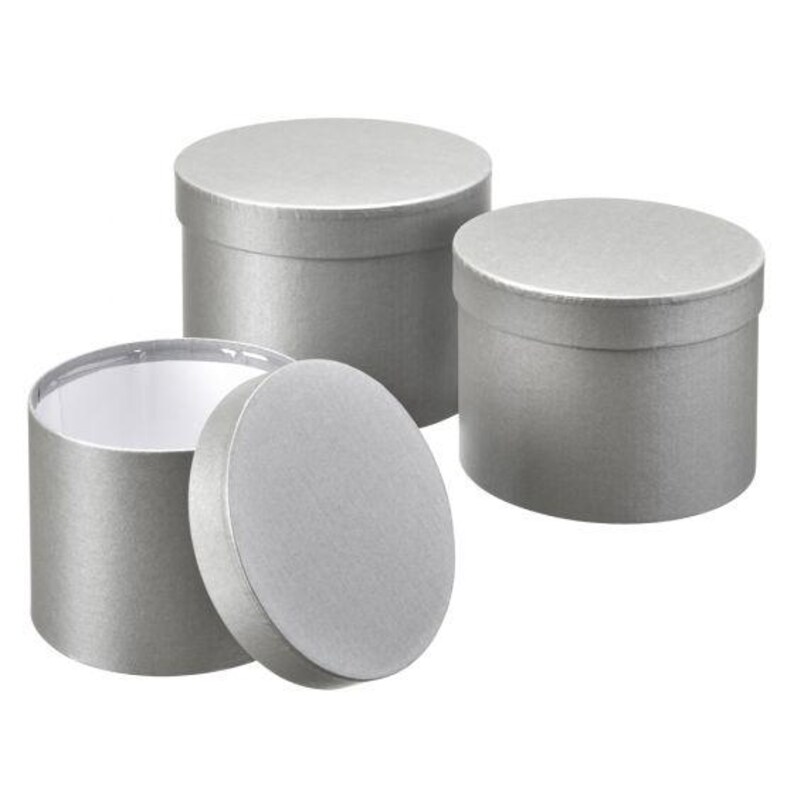 May include: Three cylindrical gift boxes with a metallic silver finish. Two boxes are closed with matching lids, while one box is open with its circular lid placed upright in front of it, revealing a plain white interior lining.