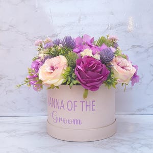 May include: A cylindrical light pink gift box featuring the purple text NANNA OF THE GROOM. The box is filled with an arrangement of artificial flowers including deep purple roses, light pink roses, purple thistles, and small green foliage.