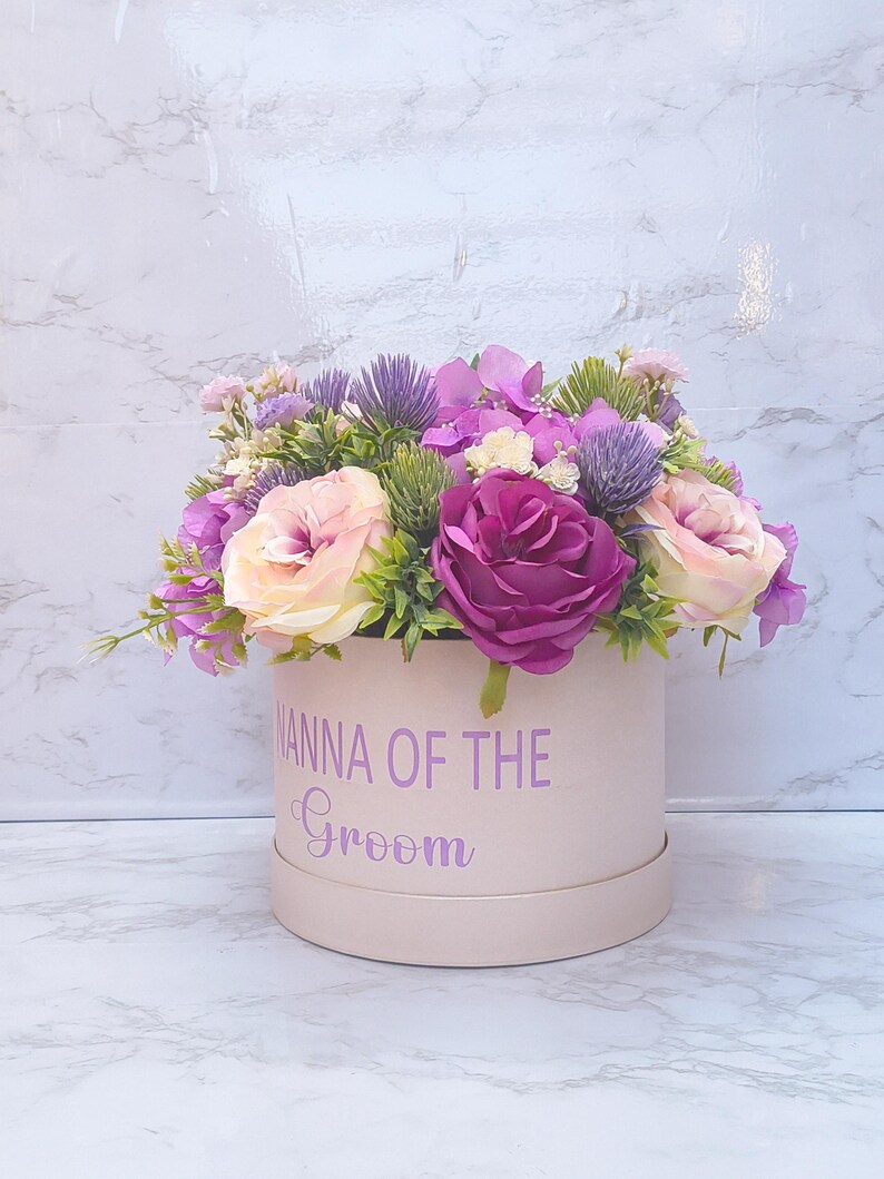 May include: A cylindrical light pink gift box featuring the purple text NANNA OF THE GROOM. The box is filled with an arrangement of artificial flowers including deep purple roses, light pink roses, purple thistles, and small green foliage.
