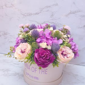 May include: A round light pink gift box featuring the purple text "MOMMA OF THE GROOM" filled with an arrangement of artificial flowers including deep magenta roses, pale pink roses, purple hydrangeas, spiky purple thistles, and green foliage.