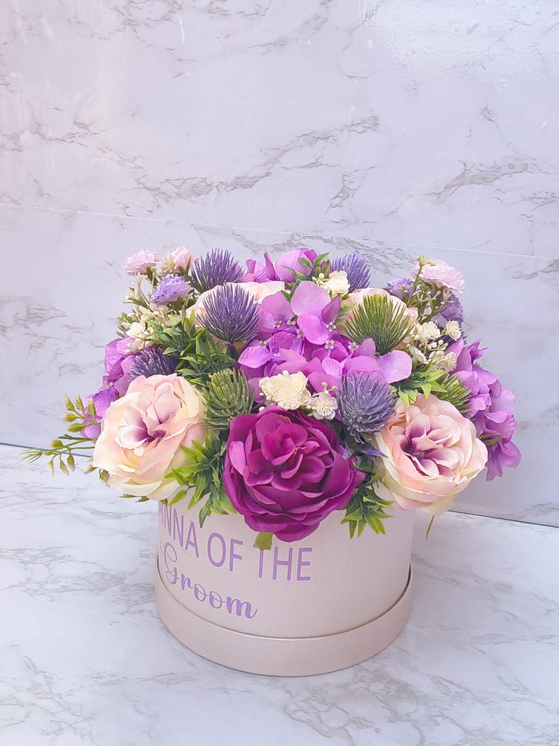 May include: A round light pink gift box featuring the purple text "MOMMA OF THE GROOM" filled with an arrangement of artificial flowers including deep magenta roses, pale pink roses, purple hydrangeas, spiky purple thistles, and green foliage.