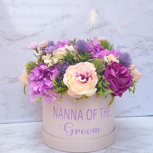 May include: A round, light pink gift box featuring the purple text "NANNA OF THE Groom" on the front, filled with an arrangement of artificial flowers including purple hydrangeas, light pink roses, and spiky purple thistle-like accents.