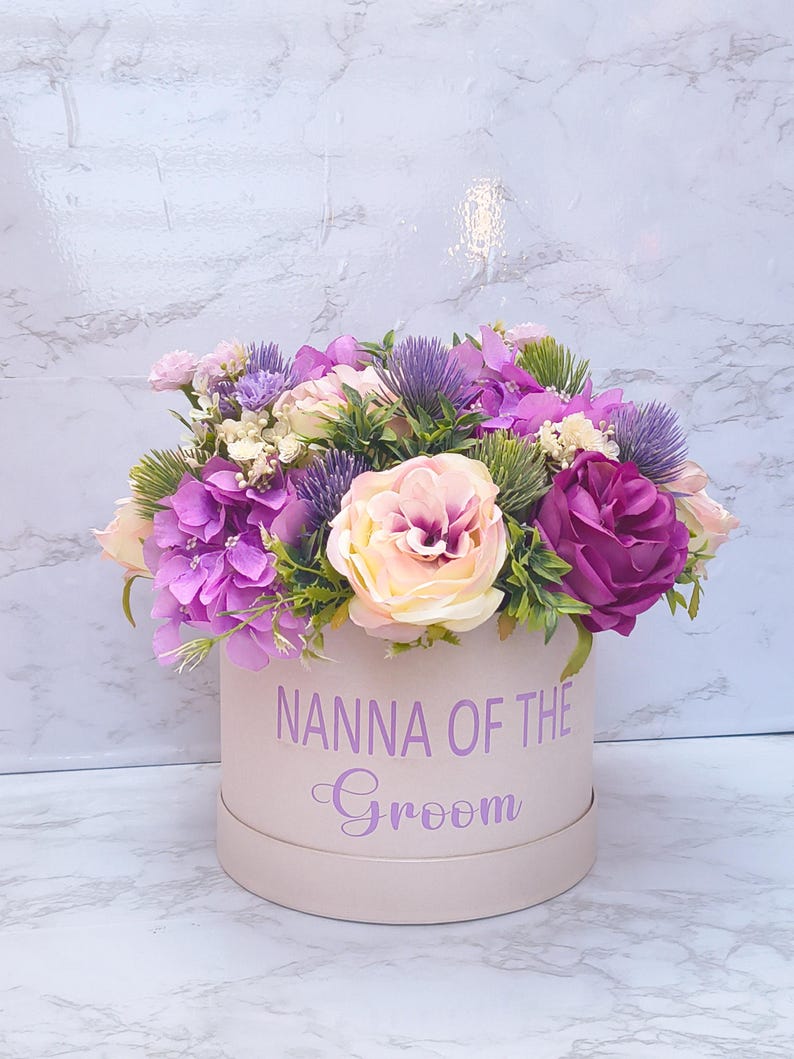May include: A round, light pink gift box featuring the purple text "NANNA OF THE Groom" on the front, filled with an arrangement of artificial flowers including purple hydrangeas, light pink roses, and spiky purple thistle-like accents.