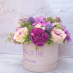 May include: A round, light pink gift box featuring the purple text "ANNA OF THE Groom" on the front, filled with an arrangement of artificial flowers including deep magenta roses, pale pink roses, purple thistle, and small purple hydrangea.