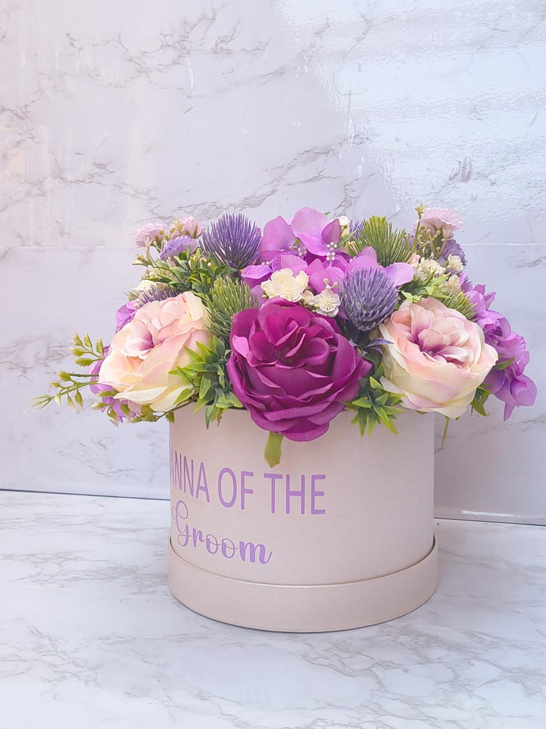 May include: A round, light pink gift box featuring the purple text "ANNA OF THE Groom" on the front, filled with an arrangement of artificial flowers including deep magenta roses, pale pink roses, purple thistle, and small purple hydrangea.