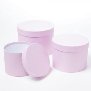 May include: A set of three cylindrical light pink gift boxes with matching lids. One box is shown open with its lid placed in front, while the other two boxes remain closed. The interior of the open box is white, and the exterior is a solid matte light pink.