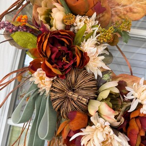 May include: A decorative wreath featuring deep red and orange faux peonies, cream-colored chrysanthemums, and a central woven twig pumpkin. The arrangement includes dried brown leaves, green foliage, small purple berries, and a sage green velvet ribbon.