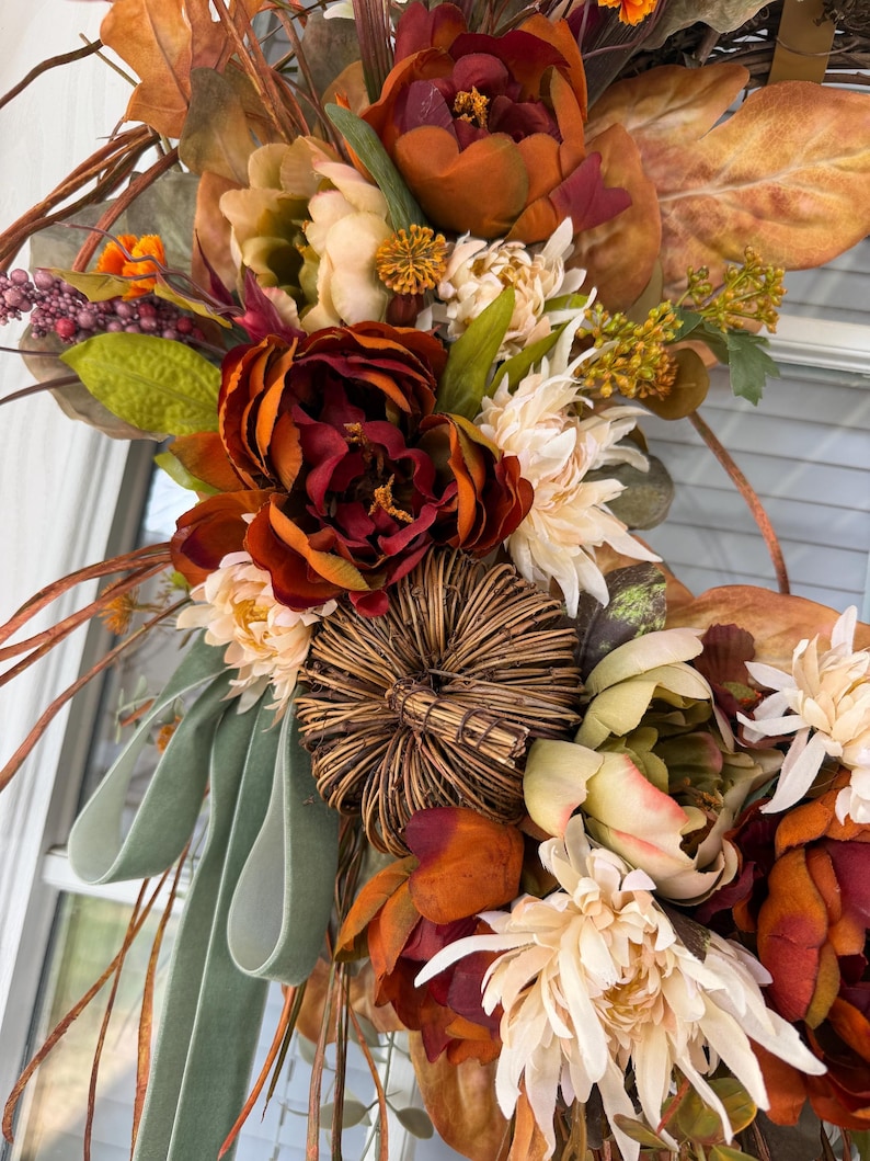 May include: A decorative wreath featuring deep red and orange faux peonies, cream-colored chrysanthemums, and a central woven twig pumpkin. The arrangement includes dried brown leaves, green foliage, small purple berries, and a sage green velvet ribbon.