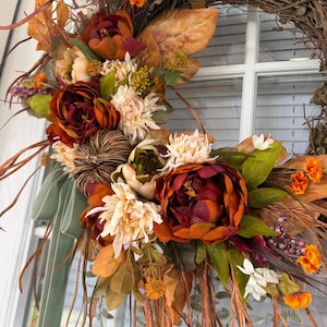 May include: A decorative wreath featuring a grapevine base adorned with artificial autumn foliage, including deep red and orange peonies, cream-colored chrysanthemums, small orange marigolds, a brown woven pumpkin, and long sage green velvet ribbons.