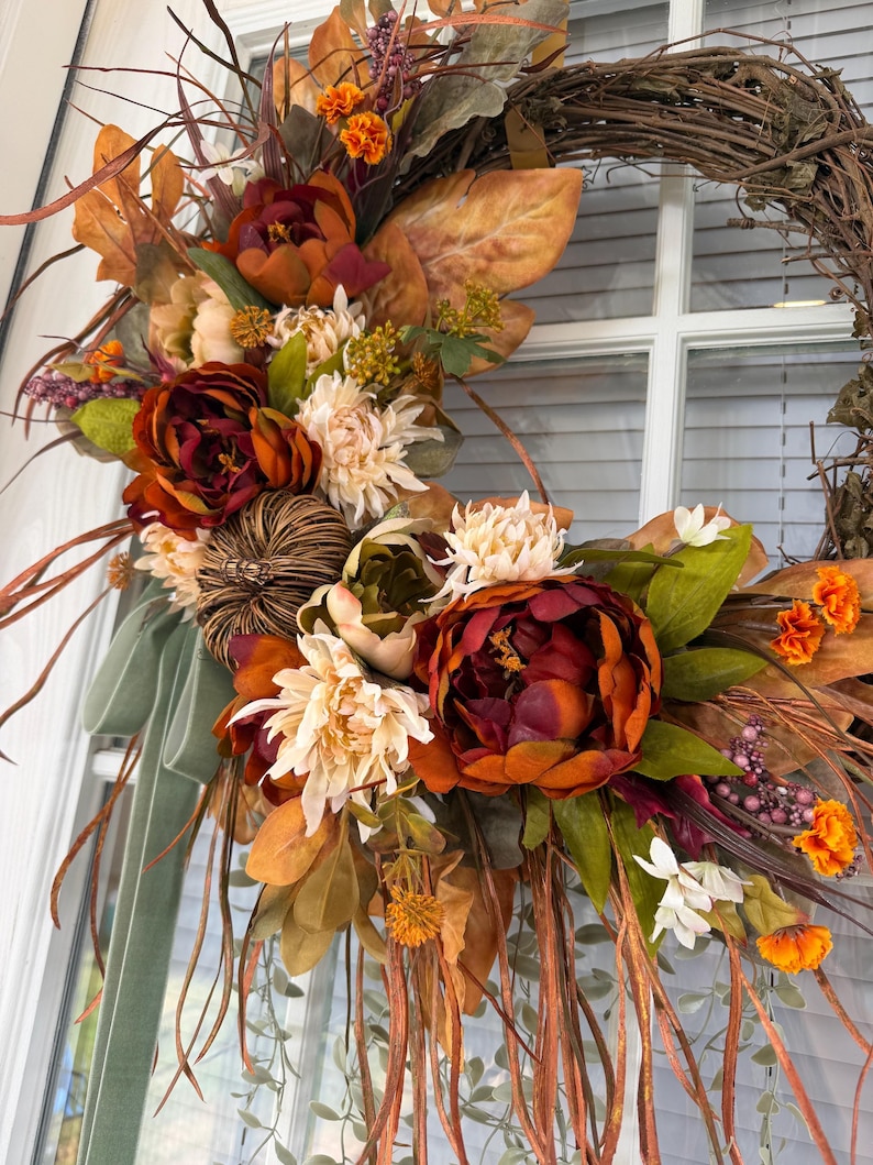 May include: A decorative wreath featuring a grapevine base adorned with artificial autumn foliage, including deep red and orange peonies, cream-colored chrysanthemums, small orange marigolds, a brown woven pumpkin, and long sage green velvet ribbons.