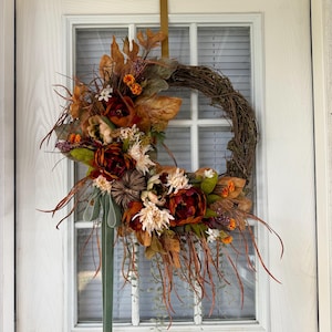 May include: A circular grapevine wreath adorned with artificial autumn foliage, including deep red and orange flowers, dried leaves, small pumpkins, and wispy brown grasses. A long, sage green ribbon hangs from the bottom, and a gold metal hook is attached.