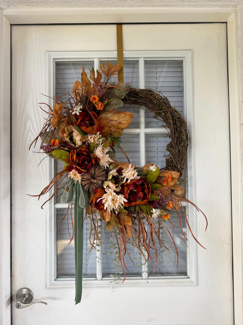 May include: A circular grapevine wreath adorned with artificial autumn foliage, including deep red and orange flowers, dried leaves, small pumpkins, and wispy brown grasses. A long, sage green ribbon hangs from the bottom, and a gold metal hook is attached.