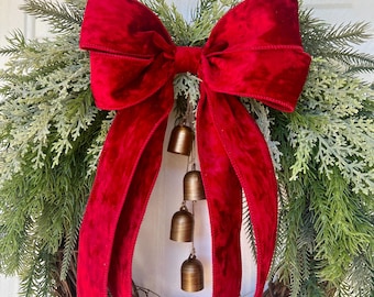 May include: A wreath constructed from brown twigs and green pine needles, featuring a large, bright red velvet bow at the top center. Four small, metallic bronze-colored bells hang vertically from the center of the bow on thin strings.