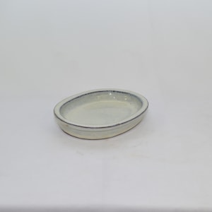 May include: A white oval ceramic dish with a blue rim. The dish is slightly raised in the center and has a smooth, glazed surface.