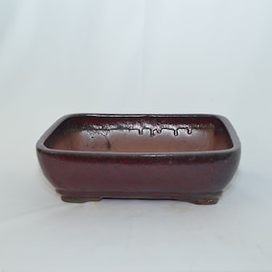 May include: A square, red-brown ceramic bonsai pot with a textured surface. The pot has a slightly rounded bottom and four small feet.