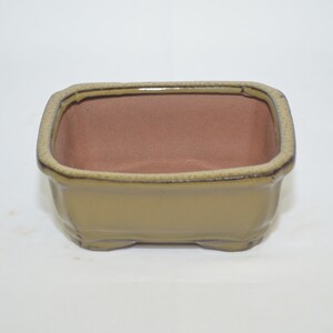 May include: A square, light green ceramic bonsai pot with a brown interior. The pot has a slightly raised rim and four small feet.