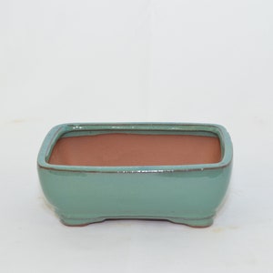 May include: A rectangular, light green ceramic bonsai pot with a brown clay interior. The pot has a slightly rounded bottom and four small feet.