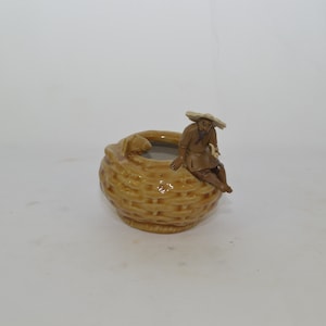 May include: A small ceramic planter shaped like a woven basket with a figurine of a man wearing a straw hat sitting on top. The planter is brown and the figurine is brown and beige.