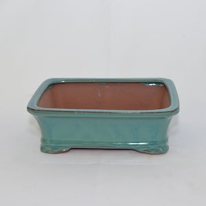 May include: A rectangular bonsai pot with a light blue glaze and a brown clay interior. The pot has four small feet.