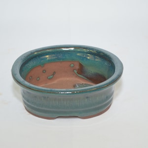 May include: A teal glazed oval bonsai pot with a brown clay base. The pot has a slightly raised rim and a textured surface.